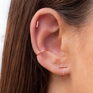 May include: A person's ear with three gold earrings. The top earring is a small bar, the middle earring is a thin hoop, and the bottom earring is a bar with a small hook.