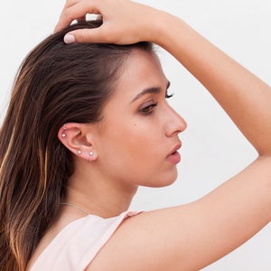 May include: A woman with long brown hair is wearing multiple small silver earrings in her left ear. She is wearing a light pink sleeveless top.