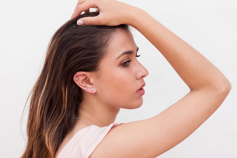 May include: A woman with long brown hair is wearing multiple small silver earrings in her left ear. She is wearing a light pink sleeveless top.