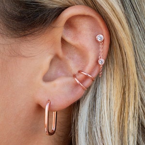 May include: A close-up of a person's ear with multiple earrings. The earrings are made of rose gold and feature a variety of styles, including a hoop, a chain with two small stones, and a double hoop.