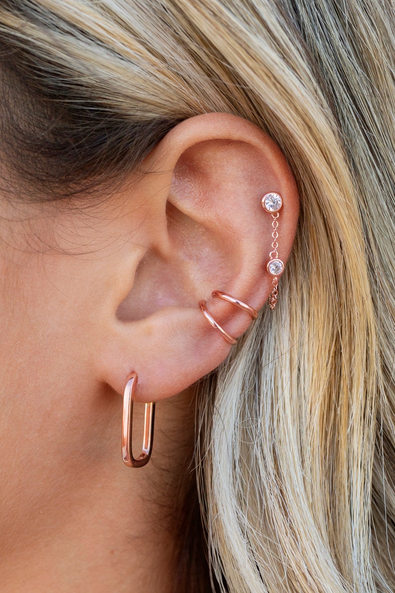 May include: A close-up of a person's ear with multiple earrings. The earrings are made of rose gold and feature a variety of styles, including a hoop, a chain with two small stones, and a double hoop.