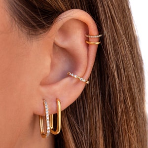 May include: Gold hoop earrings with a single row of clear cubic zirconia stones. The earrings are shown on a model's ear with other gold earrings.