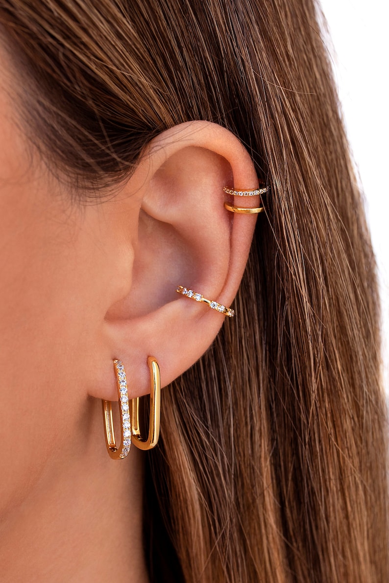 May include: Gold hoop earrings with a single row of clear cubic zirconia stones. The earrings are shown on a model's ear with other gold earrings.