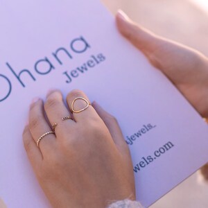 May include: A hand wearing three gold rings. The rings are a thin band with small beads, a thin band with a curved shape, and a thicker band with a larger curved shape. The hand is holding a white card with the text "Ohana Jewels" and the website address "ohana.jewels.com".