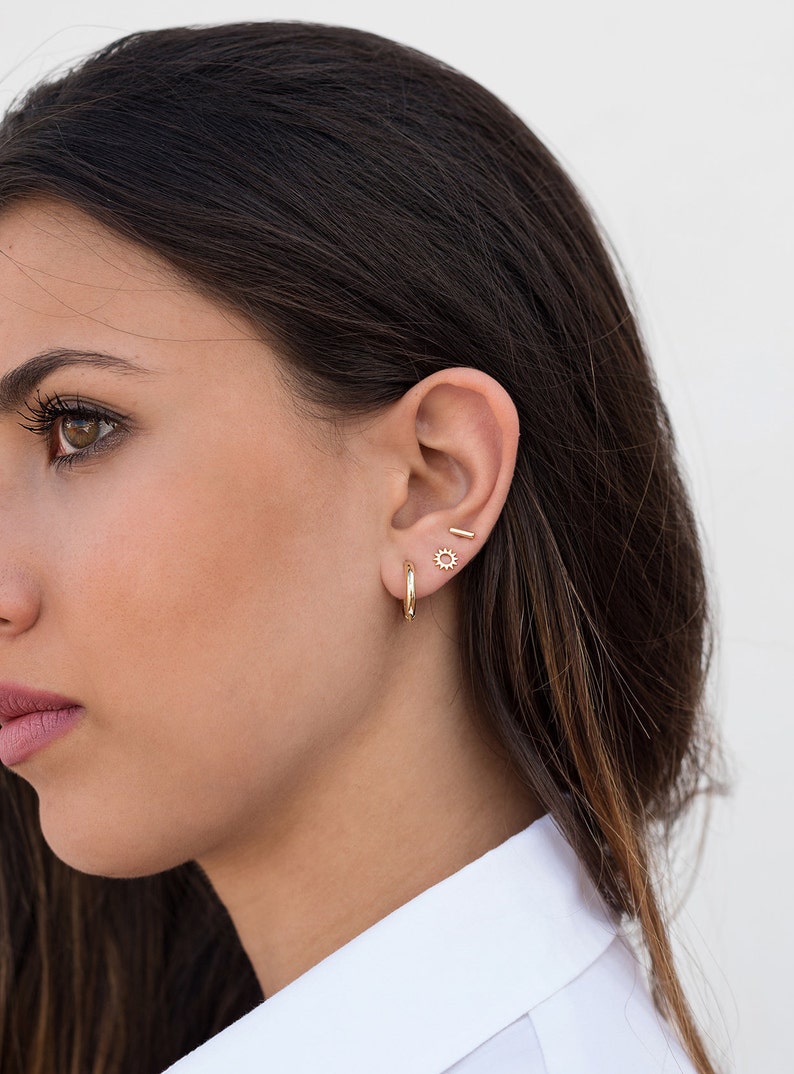 May include: A woman wearing gold hoop earrings, a small gold sun earring, and a gold bar earring.