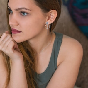 May include: A woman with long brown hair wearing a green tank top and silver earrings. She is sitting on a couch and looking off to the side.