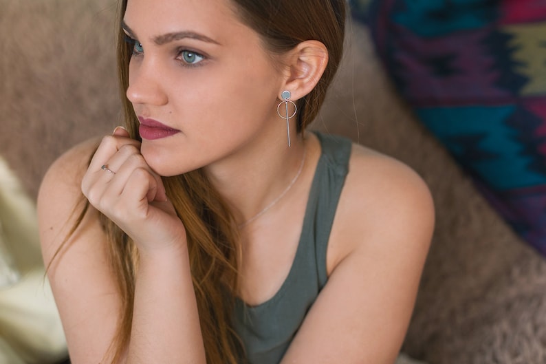 May include: A woman with long brown hair wearing a green tank top and silver earrings. She is sitting on a couch and looking off to the side.