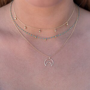 May include: A layered gold necklace with a crescent moon pendant. The necklace features two delicate chains with small turquoise beads and a thicker chain with larger gold beads.