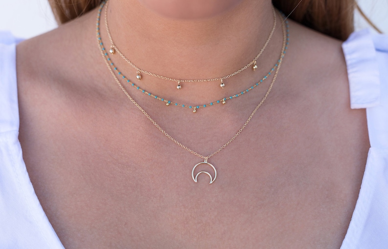 May include: A layered gold necklace with a crescent moon pendant. The necklace features two delicate chains with small turquoise beads and a thicker chain with larger gold beads.