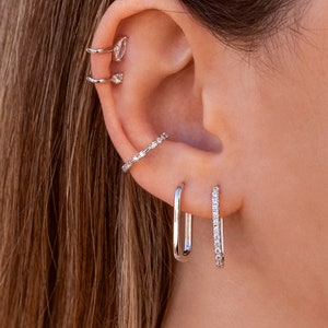 May include: A close-up of a person's ear with multiple silver earrings. The earrings are all different styles, including a hoop, a stud, and a curved bar. The earrings are all set with small, clear stones.