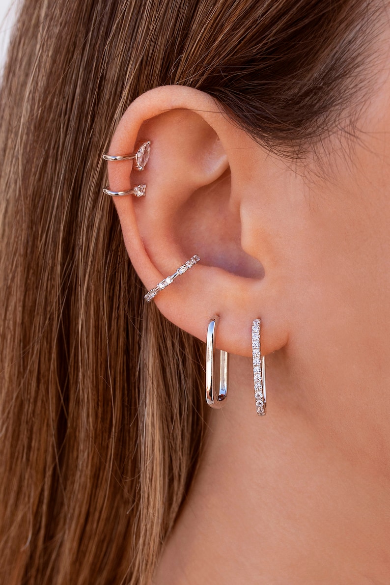 May include: A close-up of a person's ear with multiple silver earrings. The earrings are all different styles, including a hoop, a stud, and a curved bar. The earrings are all set with small, clear stones.