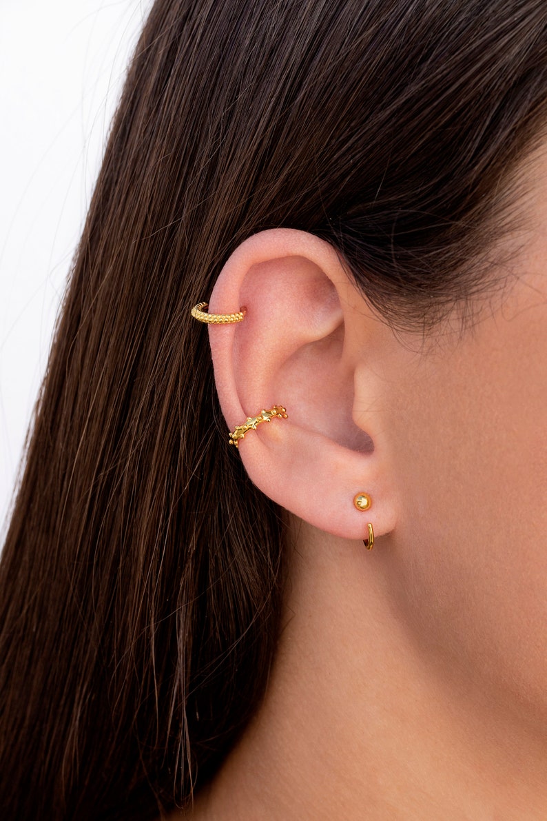 Puede incluir: Pendientes de aro de oro con un dise&ntilde;o texturizado. El aro se lleva en la parte superior de la oreja. Un peque&ntilde;o pendiente de bot&oacute;n de oro se lleva en el l&oacute;bulo inferior de la oreja. Un aro de oro se lleva en el l&oacute;bulo inferior de la oreja.