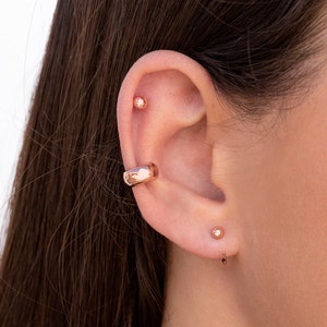 Puede incluir: Primer plano de la oreja de una persona con tres pendientes de oro. Los pendientes son un peque&ntilde;o pendiente de bot&oacute;n, un pendiente de bot&oacute;n m&aacute;s grande y un aro curvo.