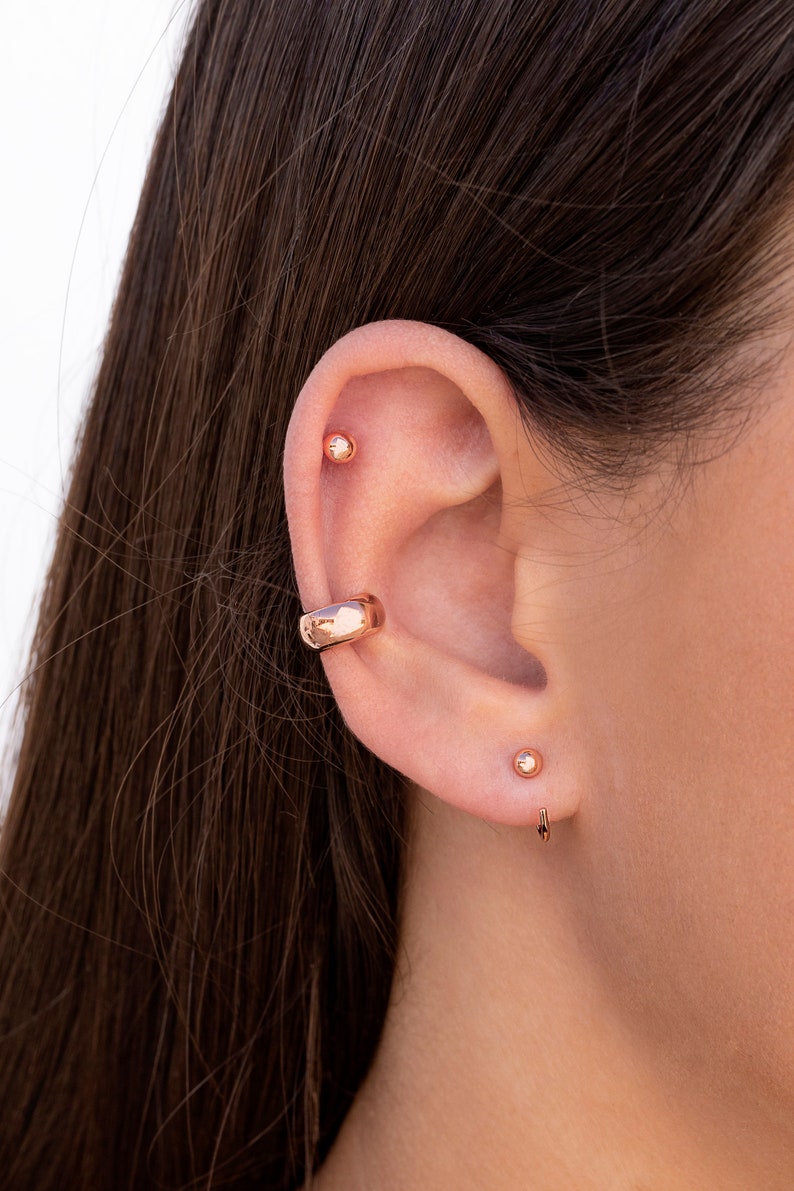 Puede incluir: Primer plano de la oreja de una persona con tres pendientes de oro. Los pendientes son un peque&ntilde;o pendiente de bot&oacute;n, un pendiente de bot&oacute;n m&aacute;s grande y un aro curvo.