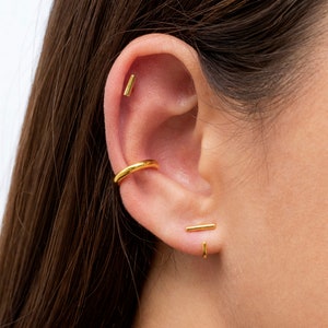 May include: A woman's ear with three gold earrings. The top earring is a small bar, the middle earring is a hoop, and the bottom earring is a bar.
