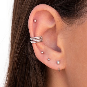 Puede incluir: Primer plano de una oreja con m&uacute;ltiples perforaciones. La oreja est&aacute; adornada con un pendiente de aro de plata, un peque&ntilde;o pendiente de bot&oacute;n de plata y un aro de plata.