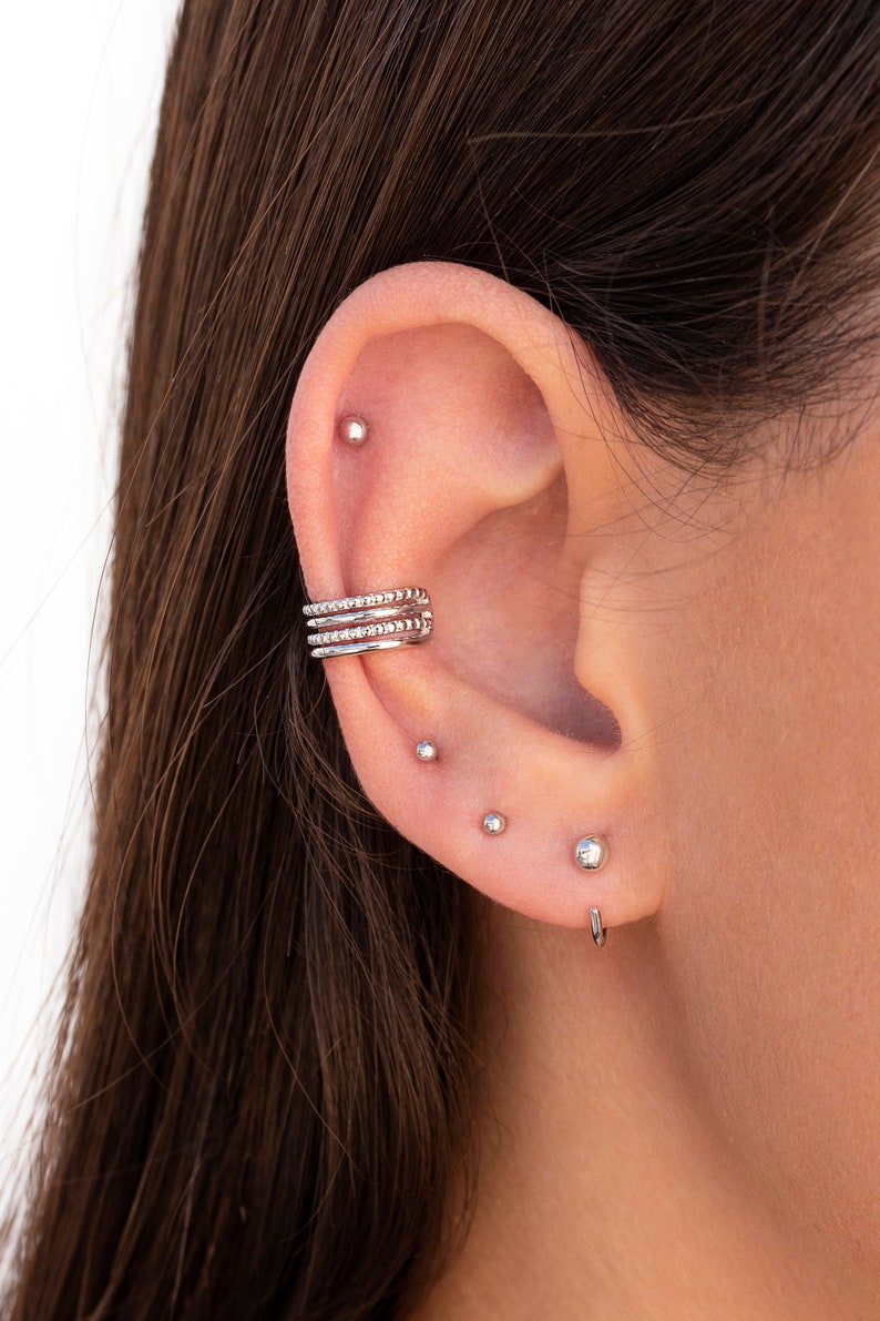 Puede incluir: Primer plano de una oreja con m&uacute;ltiples perforaciones. La oreja est&aacute; adornada con un pendiente de aro de plata, un peque&ntilde;o pendiente de bot&oacute;n de plata y un aro de plata.