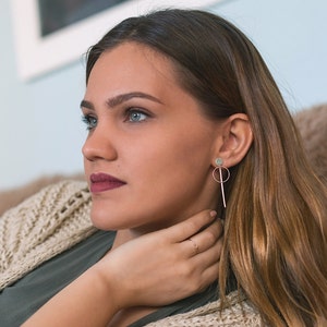 May include: A woman with long brown hair wearing a green shirt and a beige cardigan. She is wearing a pair of gold earrings with a small circle and a long, thin rod.
