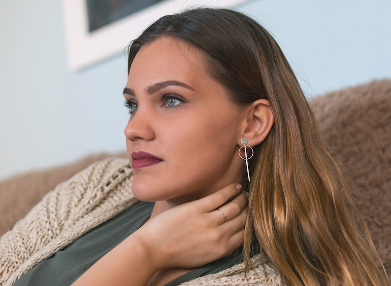 May include: A woman with long brown hair wearing a green shirt and a beige cardigan. She is wearing a pair of gold earrings with a small circle and a long, thin rod.
