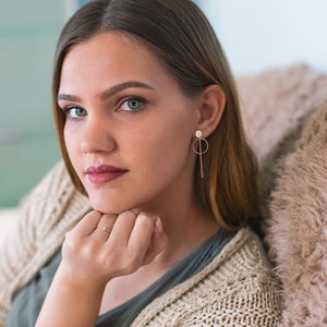 May include: A young woman with long brown hair wearing a beige knitted cardigan and a green shirt. She is wearing a pair of gold earrings with a small white pearl and a long, thin gold bar hanging from each earring. She is looking at the camera with a serious expression.