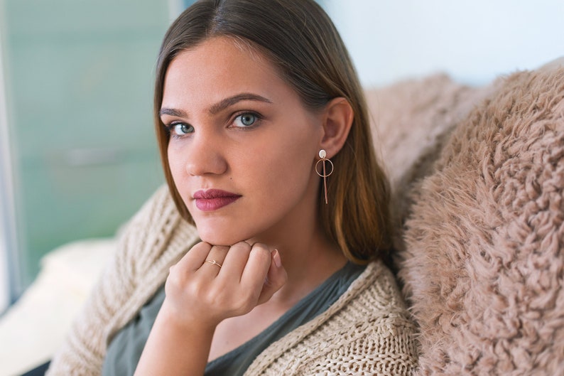 May include: A young woman with long brown hair wearing a beige knitted cardigan and a green shirt. She is wearing a pair of gold earrings with a small white pearl and a long, thin gold bar hanging from each earring. She is looking at the camera with a serious expression.