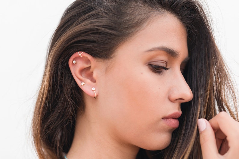 May include: A woman with long brown hair wearing multiple small gold earrings in her left ear. The earrings include a hoop, a stud, and a small bar.