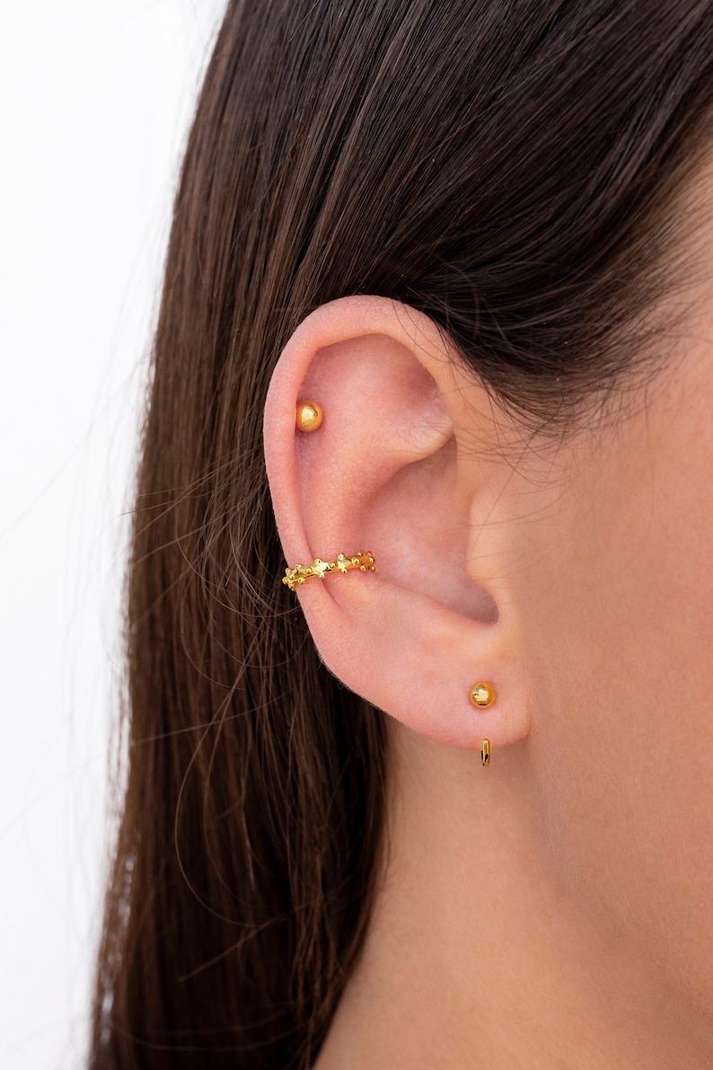 Puede incluir: Un aro de oreja dorado con un dise&ntilde;o de estrella y dos pendientes de bot&oacute;n dorados. El aro de oreja se lleva en la parte superior de la oreja y los pendientes de bot&oacute;n se llevan en la parte inferior de la oreja.