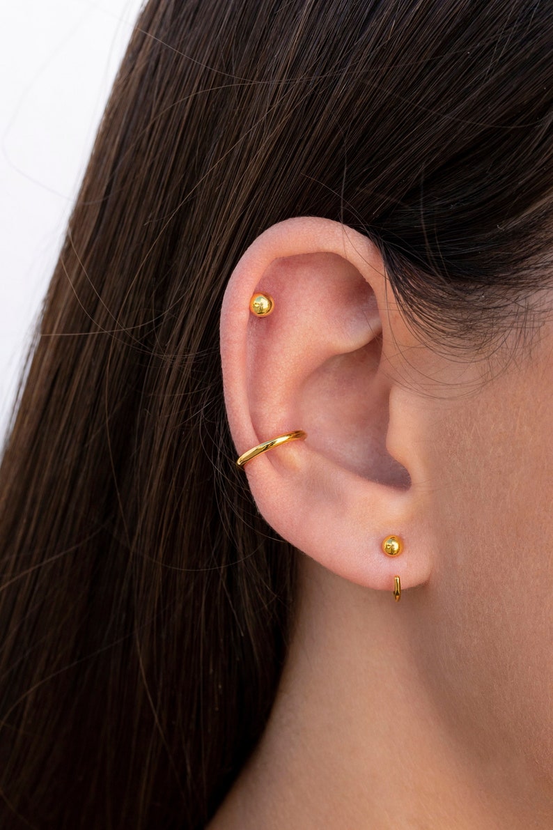 Puede incluir: Primer plano de la oreja de una persona con tres pendientes de oro. Los pendientes son un peque&ntilde;o pendiente de bola, un aro y un peque&ntilde;o pendiente de bola con una barra curva.
