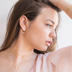 May include: A woman wearing a delicate gold necklace with a small circle pendant and a matching pair of small hoop earrings. She is wearing a light pink top and has her hair pulled back.