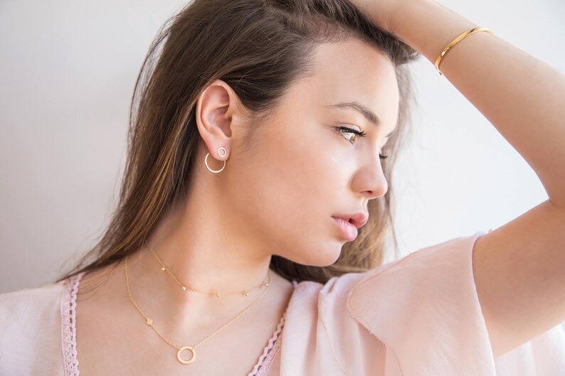 May include: A woman wearing a delicate gold necklace with a small circle pendant and a matching pair of small hoop earrings. She is wearing a light pink top and has her hair pulled back.