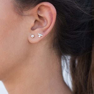 May include: A close-up of a person's ear with two small earrings. The earring on the lower lobe is a small round stone, and the earring on the upper lobe is a thin, horizontal bar.