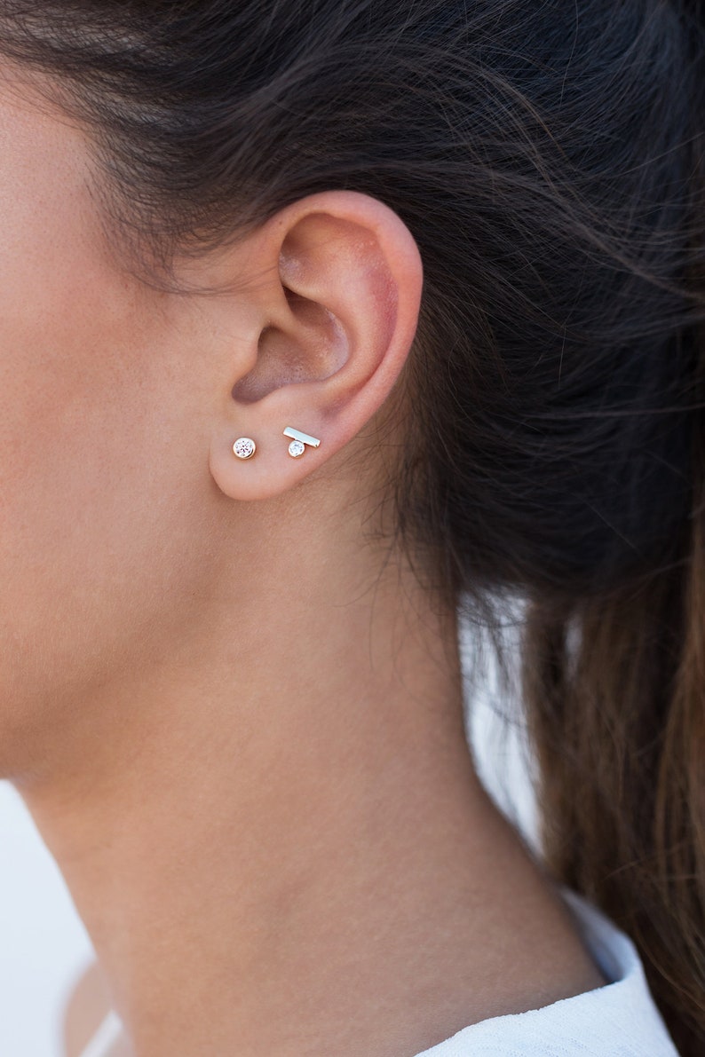 May include: A close-up of a person's ear with two small earrings. The earring on the lower lobe is a small round stone, and the earring on the upper lobe is a thin, horizontal bar.