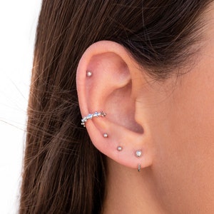 Puede incluir: Primer plano de la oreja de una persona con varios pendientes de plata. Los pendientes incluyen un peque&ntilde;o pendiente de bot&oacute;n, un pendiente de bot&oacute;n m&aacute;s grande y un pendiente de aro curvo con un delicado dise&ntilde;o de cadena.