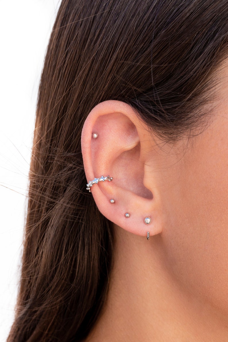 Puede incluir: Primer plano de la oreja de una persona con varios pendientes de plata. Los pendientes incluyen un peque&ntilde;o pendiente de bot&oacute;n, un pendiente de bot&oacute;n m&aacute;s grande y un pendiente de aro curvo con un delicado dise&ntilde;o de cadena.