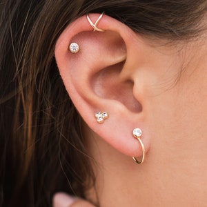 May include: A close-up of a person's ear with multiple gold earrings. The earrings feature small, clear gemstones. One earring is a hoop with a single gemstone, another is a cluster of three gemstones, and the third is a single gemstone stud.