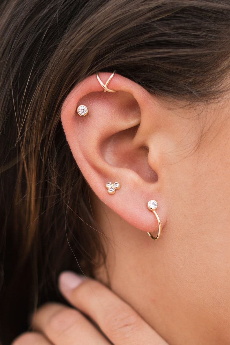 May include: A close-up of a person's ear with multiple gold earrings. The earrings feature small, clear gemstones. One earring is a hoop with a single gemstone, another is a cluster of three gemstones, and the third is a single gemstone stud.