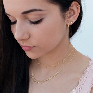 May include: A woman with long dark hair wearing a delicate gold necklace with small dangling balls and a pair of gold earrings. The necklace is layered with a thinner chain. The woman is wearing a pink top with lace trim.