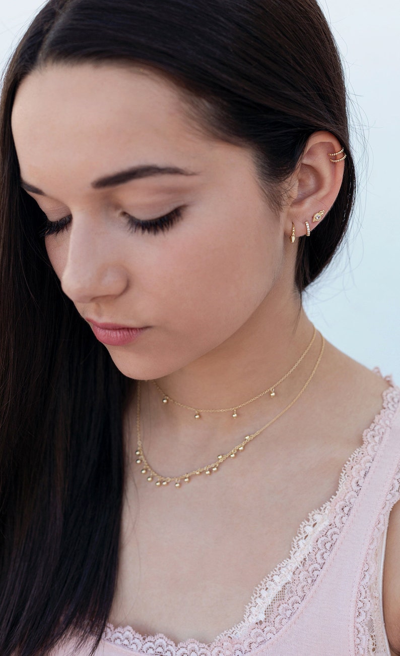 May include: A woman with long dark hair wearing a delicate gold necklace with small dangling balls and a pair of gold earrings. The necklace is layered with a thinner chain. The woman is wearing a pink top with lace trim.