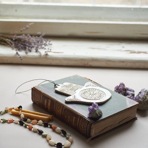 May include: A silver hamsa hand charm with intricate details sits on top of a vintage green book with a purple crystal next to it. A beaded necklace with colorful stones and two yellow candles are in the foreground.