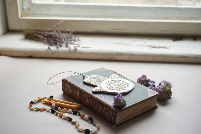 May include: A silver hamsa hand charm with intricate details sits on top of a vintage green book with a purple crystal next to it. A beaded necklace with colorful stones and two yellow candles are in the foreground.