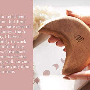 May include: A hand holds a crescent moon-shaped wooden sculpture. The sculpture is light brown and has a small, embossed logo that reads "bil glyne".