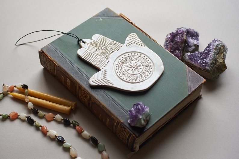 May include: White ceramic hamsa hand with intricate details, hanging from a string, on top of a green book with a purple crystal next to it.
