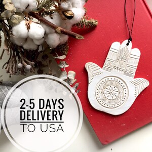 May include: White ceramic Hamsa hand ornament with intricate details, hanging from a black string. The ornament is on a red surface with the text "2-5 DAYS DELIVERY TO USA".