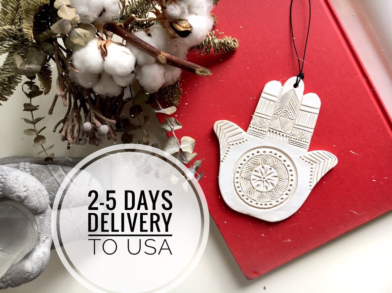 May include: White ceramic Hamsa hand ornament with intricate details, hanging from a black string. The ornament is on a red surface with the text "2-5 DAYS DELIVERY TO USA".