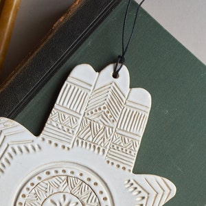 May include: White ceramic hand-shaped ornament with an intricate embossed design. The ornament is hanging from a black string.