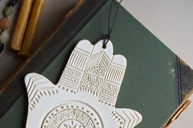 May include: White ceramic hand-shaped ornament with an intricate embossed design. The ornament is hanging from a black string.