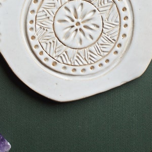May include: White ceramic dish with a raised, circular design. The design features a central flower with a ring of geometric patterns around it. The dish is on a dark green surface.