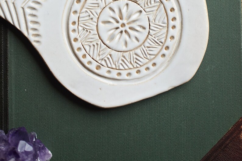 May include: White ceramic dish with a raised, circular design. The design features a central flower with a ring of geometric patterns around it. The dish is on a dark green surface.