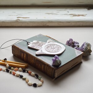 May include: A vintage book with a green cover and a white ceramic hamsa charm hanging from it. The book is surrounded by purple amethyst crystals and two yellow candles.