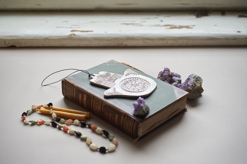 May include: A vintage book with a green cover and a white ceramic hamsa charm hanging from it. The book is surrounded by purple amethyst crystals and two yellow candles.